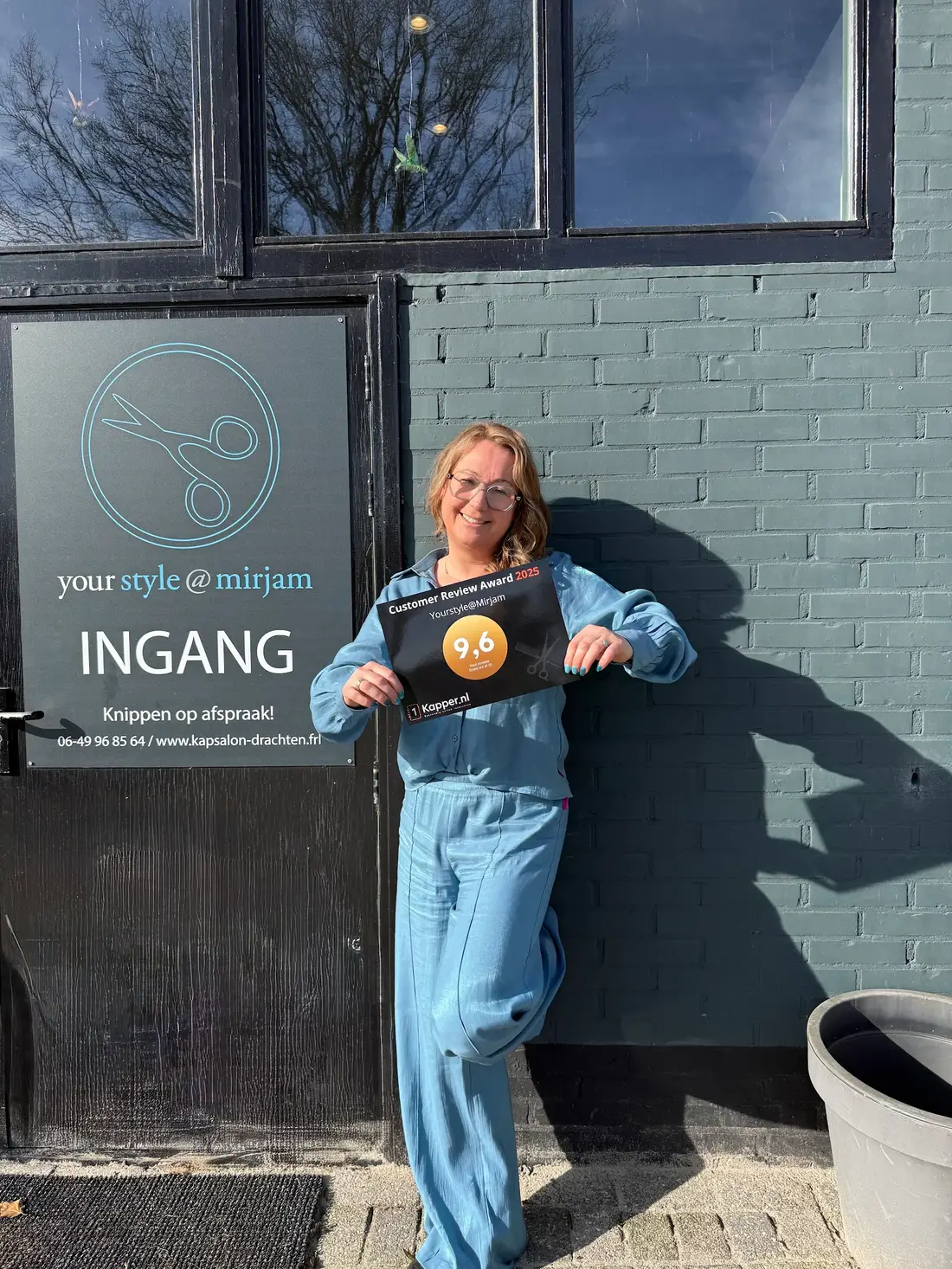 Mirjam staat lachend tegen de groene gevel naast de ingang van haar kapsalon